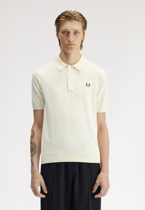 Jeune homme aux cheveux courts portant un polo crème à manches courtes et un pantalon noir, debout devant un fond clair uni.