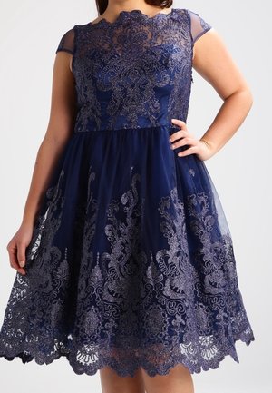 Tmavě modré šaty délky po kolena s jemnou krajkovou vrstvou, průsvitnými krátkými rukávy a květinovou výšivkou, nošené osobou s rukou na boku.