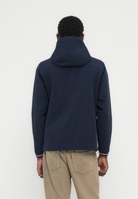 Άνδρας που φοράει ναυτικό μπλε μπουφάν με κουκούλα και μπεζ τζιν, στέκεται στραμμένος σε έναν απλό ανοιχτόχρωμο τοίχο.