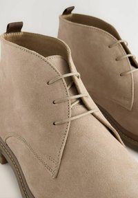 Beige Wildleder-Ankle Boots mit glatter Textur, runden Zehen, niedrigem Absatz und einfacher Schnürung. Enthält eine Schlaufe an der Rückseite.