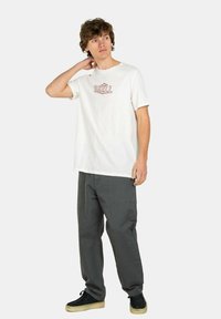 White cotton t-shirt with red "REELL" graphic, paired with loose gray pants and black sneakers. Simple design, casual fit, minimal texture.