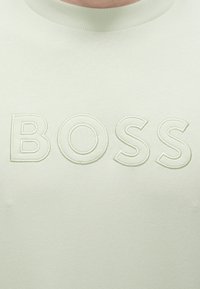 Gros plan d'une chemise beige clair avec des lettres brodées en relief formant le mot BOSS sur la poitrine sous le col.
