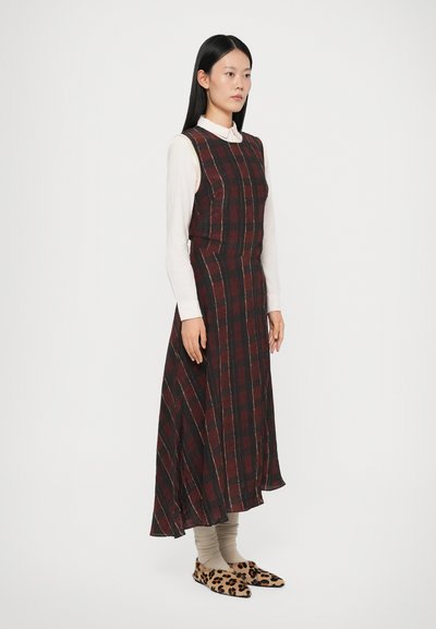 Abito lungo a quadri in marrone, rosso e nero con un bustino aderente e una gonna svasata. Indossato sopra una camicia bianca e abbinato a scarpe con fantasia.