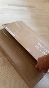 Roka z ročno zapestnico odpirajo veliko, delno odprto kartonsko škatlo Zalando na svetlem lesenem tleh.