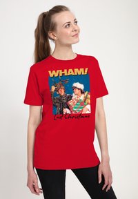 Kobieta ubrana w czerwoną koszulkę z grafiką Wham! Last Christmas, na której widać dwóch mężczyzn - jednego w kostiumie renifera, a drugiego w stroju Świętego Mikołaja, trzymających prezenty.