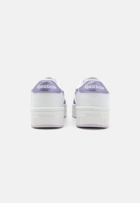 Bela tenisa z vijoličnimi detajli. Gladka usnjena zgornji del, oblazinjen petni del, teksturiran podplat in "Reebok" oznaka na hrbtni strani. Masivna zasnova.