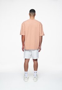 Světle broskvová bavlněná tričko s krátkým rukávem, v kombinaci s bílými šortkami. Model nosí bílé tenisky a ponožky po kotníky. Jednoduchý design.