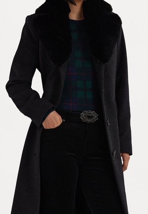 Personne portant un manteau noir avec un large col en fourrure, une chemise à carreaux verts et bleus, un pantalon noir, et une boucle de ceinture décorative, main dans la poche.