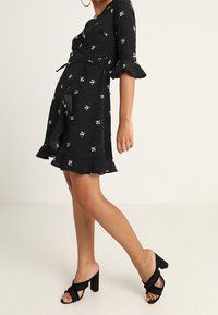 Schwarzes Kleid mit weißem Blumenmuster, Rüschensäumen und Taillenknoten. Kombiniert mit schwarzen Blockabsatzsandalen. Glatter Stoff, knielang.
