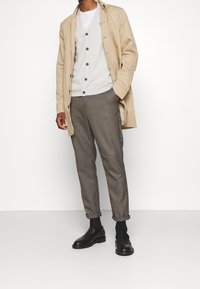 Beige übergroßes Hemd, graue Strickjacke mit Knöpfen und gemusterte braune Hose. Schwarze Mokassins runden das Outfit ab. Texturierte Stoffe sind sichtbar.