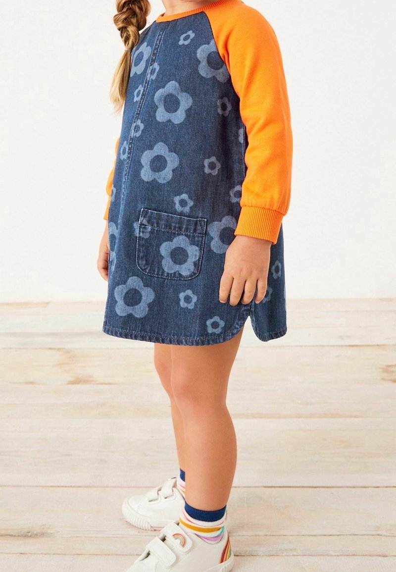 Enfant portant une robe en denim bleu avec des fleurs bleu clair, des manches longues orange, des chaussures blanches avec des motifs arc-en-ciel et des chaussettes rayées.