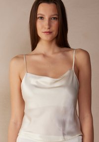 Satinkamelie in sanftem Creme mit dünnen verstellbaren Trägern und einem lockeren Cowl-Ausschnitt; glatte Textur mit minimaler Detailierung.