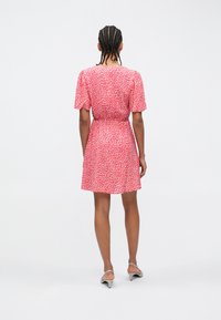 Άτομο με πλεξούδες που φοράει ένα κοντό ρούχο ροζ με λευκά στίγματα και μπεζ πέδιλα με τακούνι, στέκεται γυρισμένο προς τα πίσω μπροστά σε λευκό φόντο.