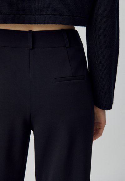 Donna che indossa pantaloni neri a vita alta e su misura con una tasca a filetto posteriore e un maglione nero a maniche lunghe cropped.