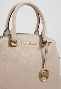 Sac à main en cuir beige texturé avec deux poignées arrondies, logo Michael Kors en or et pendentif circulaire doré MK suspendu.