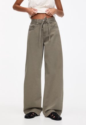 Femme portant un pantalon large vert olive avec une taille nouée et des sandales noires, ajustant son top blanc sans manches.