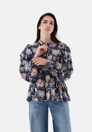 Femme portant un chemisier bleu marine à fleurs avec des manches bouffantes et un jean bleu, ajustant un poignet sur un fond neutre.