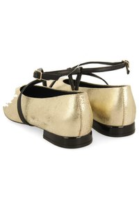 Bailarinas de cuero dorado agrietado con una correa negra en el tobillo y un bajo tacón negro. Bordes expuestos y detalle de superficie texturizada.