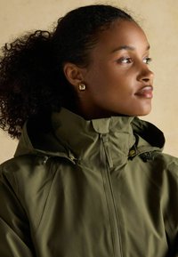 Jachetă verde impermeabilă cu guler înalt și închidere cu fermoar. Dispune de detalii din hardware auriu și o finisare netedă, texturată.