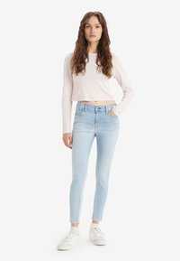 Un haut rose clair à manches longues coupé, associé à un jean skinny bleu clair. Le modèle porte des baskets blanches. Design simple, sans motifs visibles.