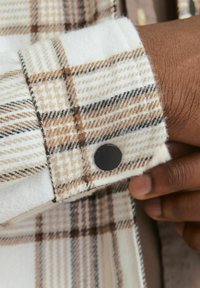 Gecarbonneerde flanellen manchetten met beige, witte en bruine patronen. Zwarte drukknoopaccent nabij de rand; zachte, gestructureerde stof.
