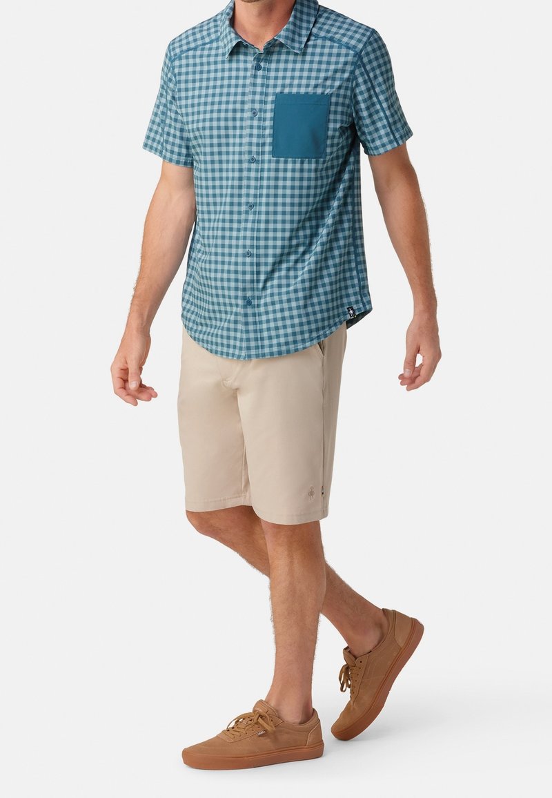 Hombre que lleva una camisa de cuadros azul de manga corta con un bolsillo verde aguamarina, pantalones cortos beiges y zapatillas marrones, caminando frente a un fondo blanco.