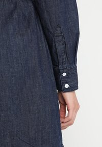 Jeansskjorta i en mörkblå nyans, med en knappförsedd manschett och synlig sömnad. Tyget har en något strukturerad yta.