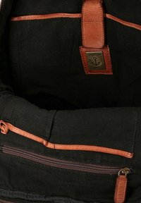 Sezione di uno zaino in tessuto nero con rifiniture in pelle marrone, una tasca con zipper e un'etichetta metallica che mostra un'ancora e la scritta "HARBOUR 2nd".