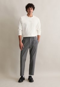 Witte lange mouwen crewneck shirt met print, gecombineerd met grijze geruite broek. Schoenen zijn zwarte instappers. Achtergrond is neutraal beige.