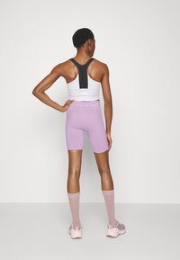Sportovní crop top v bílé barvě s černými akcenty, spárovaný se světle fialovými cyklistickými šortkami, růžovými ponožkami až po kolena a šedými sportovními botami.