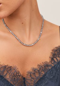 Silverfärgat halsband med pärlor som är släta, runda och fasetterade. Bärs mot en halsringning med svart spetskant.