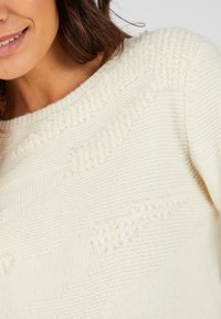 Pull en tricot couleur crème avec un motif texturé, présentant des rayures en relief et une encolure large, offrant une apparence douce et confortable.