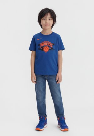 Ragazzo in piedi che indossa una maglietta blu dei New York Knicks, jeans blu e scarpe da ginnastica blu e arancioni su uno sfondo semplice.