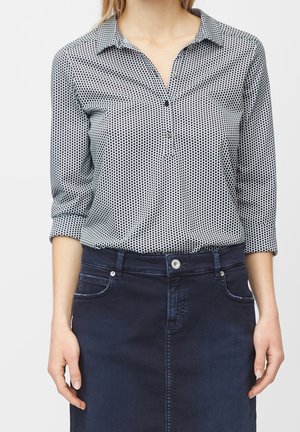 Vrouw met een witte blouse met zwarte polkadots, ingestopt in een donkerblauwe spijkerrok, met opgerolde mouwen tot de ellebogen.