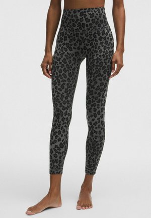 Vrouw die een high-waist legging tot op de enkel draagt met een donkergrijs en zwart luipaardprint, blootsvoets staand tegen een effen achtergrond.