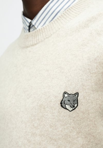 Gaiši bezsvara džemperis ar apaļu kakla izgriezumu un pelēku lapsu izšuvumu uz krūtīm. Apakšā daļēji redzams zilas un baltas svītras krekls ar apkakli.