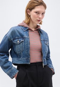 Νεαρή γυναίκα που φοράει κοντή μπλε τζιν ζακέτα πάνω από κόκκινο και άσπρο ριγέ φούτερ με κουκούλα και μαύρο ψηλόμεσο παντελόνι, με τα χέρια στις τσέπες, ουδέτερο φόντο.