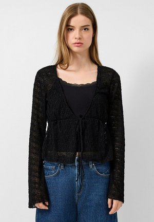 Młoda kobieta ubrana w czarną przezroczystą bluzkę z długim rękawem na czarnym topie na ramiączkach oraz niebieskie dżinsy, stoi na tle jednolitego jasnego tła.