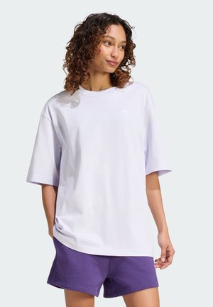 Kvinde med krøllet hår iført en løs hvid t-shirt og lilla shorts, der kigger lidt til siden med hænderne nær lommerne.