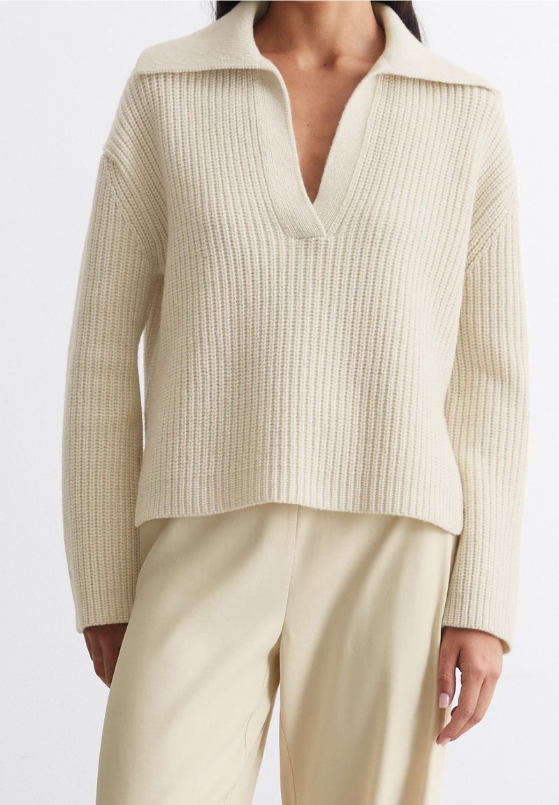 Pull tricoté de couleur crème avec un col en V et un col rabattable. Texture côtelée et coupe décontractée. Associé à un pantalon beige clair.