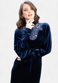 Abito di velluto blu navy con dettagli in pizzo sullo scollo e corpino aderente, dotato di maniche vaporose e fascette legate in vita.