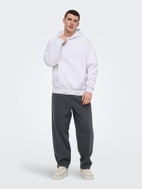 Mladi moški stoji, oblečen v belo kapucar, temno sive športne hlače in bež superge, na enobarvnem svetlo sivo ozadju.