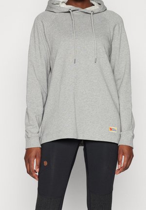 Henkilö, jolla on päällään vaaleanharmaa huppari nyöreillä ja mustat housut, joissa on paikallisia polvissa ja logo lähellä reittä.