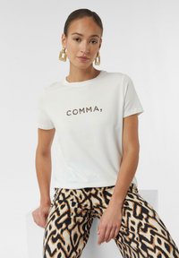 Weißes T-Shirt mit kurzen Ärmeln und dem Schriftzug "COMMA" in Leopardenmuster, kombiniert mit gemusterten beige- und schwarzen weiten Hosen.