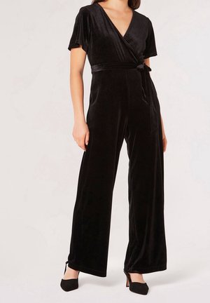 Nainen pukeutuneena mustaan samettiseen kietaisuhousupukuun, jossa on lyhyet hihat, leveät lahkeet ja solmittava vyötärönauha, yhdistetty mustiin kärjestä teräviin korkokenkiin.