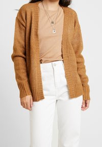 Brun stickad kofta med öppen front, ribbade muddar och lätt textur, bärs över en beige topp och vita byxor.