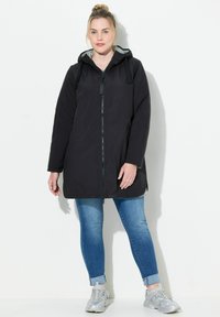 Schwarze wasserdichte Jacke mit Kapuze, durchgehendem Reißverschluss, Seitentaschen und grauem Futter, kombiniert mit blauen Jeans und silbernen Sneakers.