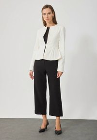 Žena nosi bijeli sako s peplum detaljima, crne široke hlače do gležnjeva i crne štikle, stojeći protiv jednostavne svijetle pozadine.