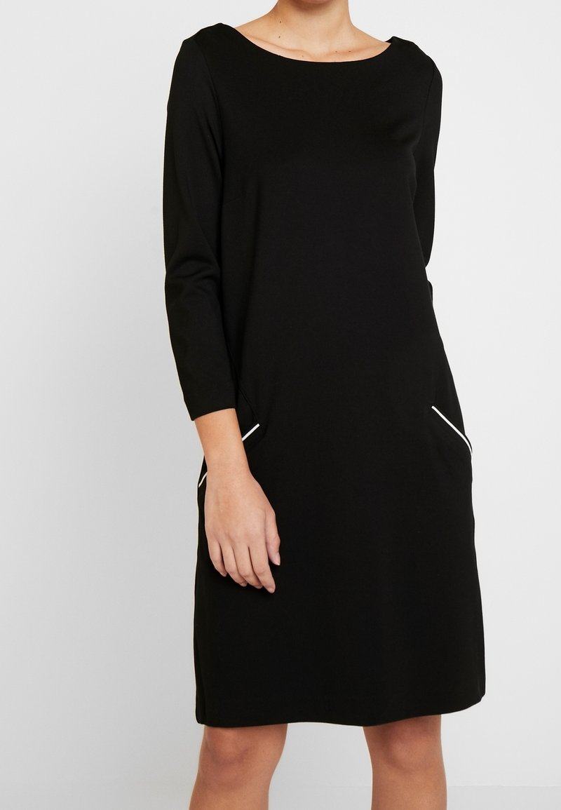 Femme portant une robe noire au genou avec des manches trois-quarts et des détails blancs sur les poches latérales, debout devant un fond uni.