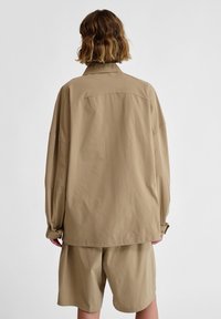 Overmaatse beige jas met een kraag, losse fit en knoopmanchetten, gecombineerd met bijpassende knielange shorts. Soepele textuur, minimalistisch ontwerp.